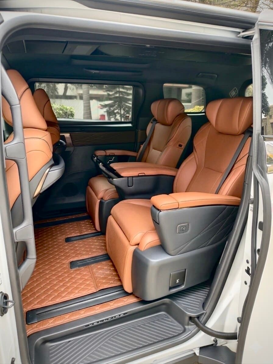 Toyota Vellfire rear cabin with cognac captain seats, fold-out tray tables, and Vellfire-embossed floor mats — spacious luxury MPV for hire in Bangalore